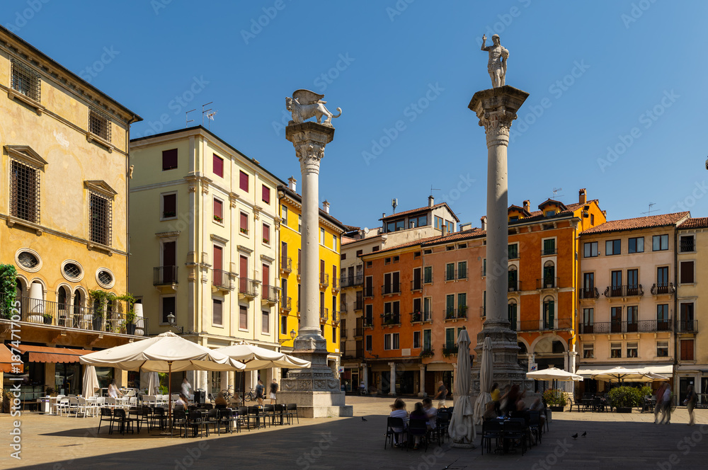 Obraz premium Two historic stone columns with sculpted figures rising in center of Piazza dei Signori in Vicenza, Italy, surrounded by colored facades of residential buildings and shaded seating areas on summer day