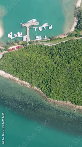 Top view of lush greenery covering a small island, turquoise waters with visible reefs, and a nearby pier with boats. Lipa Noi. Ko Samui, Thailand.
