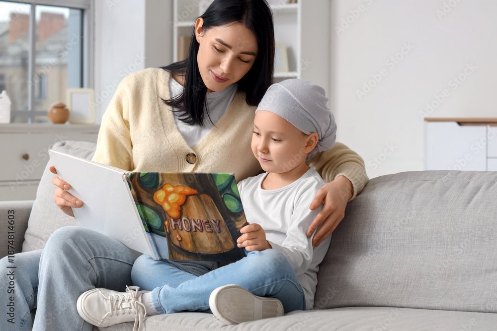 Fototapeta premium Little girl after chemotherapy with her mother reading book on sofa at home. Childhood cancer awareness concept