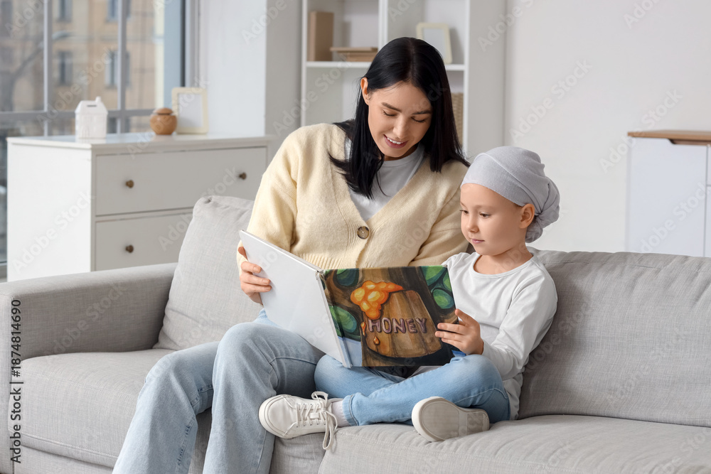 Fototapeta premium Little girl after chemotherapy with her mother reading book on sofa at home. Childhood cancer awareness concept