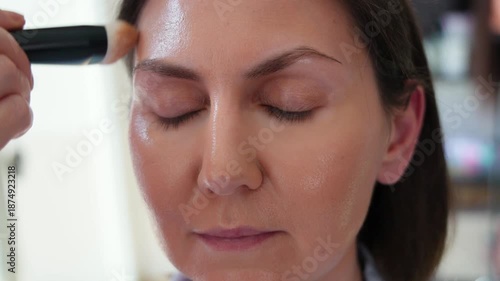 Close up of a client with eyes closed as a makeup artist applies foundation with a brush in a professional beauty salon, highlighting skincare, natural glow and modern cosmetology service