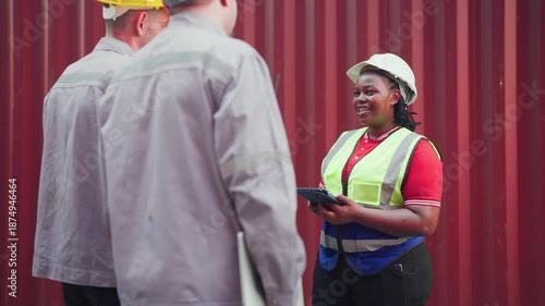 Group of diverse professional logistics managers and engineers, including an African American woman and Caucasian men, wearing safety gear and discussing work while using a digital tablet