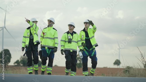 Group of professional male and female engineers in safety gear and hard hats walking through a wind turbine farm. They are conducting a site survey and project.
