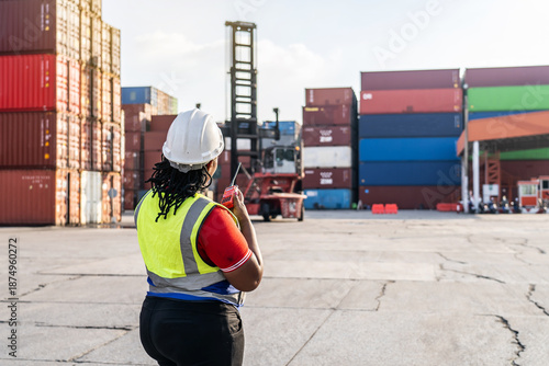 фотографія African female logistics manager in a hard hat using a walkie-talkie to coordinate container shipping operations at a busy industrial port terminal with reach stacker and cargo crates