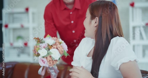 Happy young Asian man surprises his girlfriend with a colorful flower bouquet, sharing a romantic and joyful moment together in a cozy decorated living room.