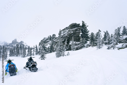 Billede på lærred Two snowmobiles parked on a snowy slope with rocky mountains and fir trees