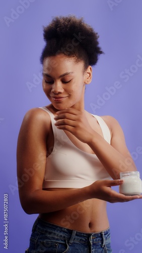Young african american woman in a laconic top takes a cream from a jar and rubs it on her shoulder while posing against a monochrome studio background, vertical video