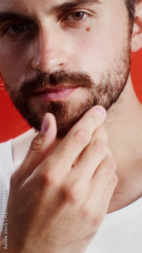 Naklejka premium Young man touching his beard while looking at camera while standing against monochrome studio background, vertical video