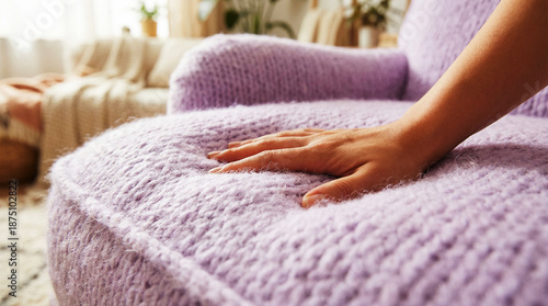A hand pressing into a soft, squishy lavender sofa with a cloud-like knitted texture. Cozy interior setting emphasizing comfort, tactile fabric, and organic furniture design.