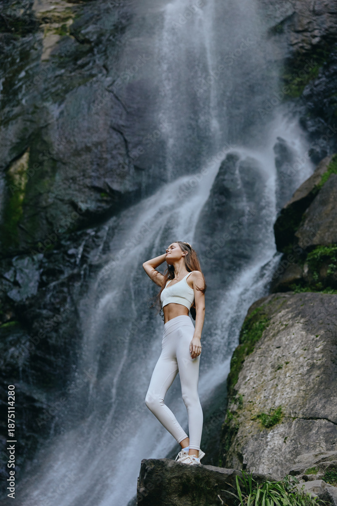 Fototapeta premium young woman standing near waterfall, wearing white sportswear, enjoying nature, serene outdoor setting, athletic and peaceful atmosphere