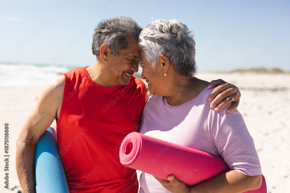 Naklejka premium Happy senior couple holding yoga mats touching foreheads of each other at beach
