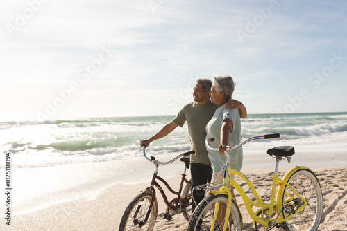 Happy multiracial senior couple looking away standing with bikes at sunny beach against sky