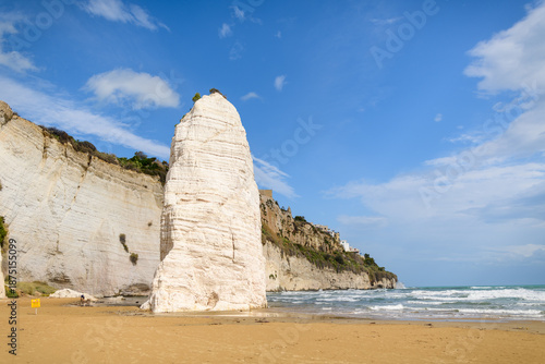 A prominent white limestone sea stack stands on the sandy beach of Vieste, Italy, framed by rugged coastal cliffs and a bright blue sky. The Adriatic Sea and distant town buildings complete this