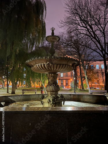 Stone fountain stands quietly in a city park at dusk, surrounded by bare trees and soft streetlight glow, Kyiv, Ukraine. The evening sky and historic architecture create a calm, timeless urban