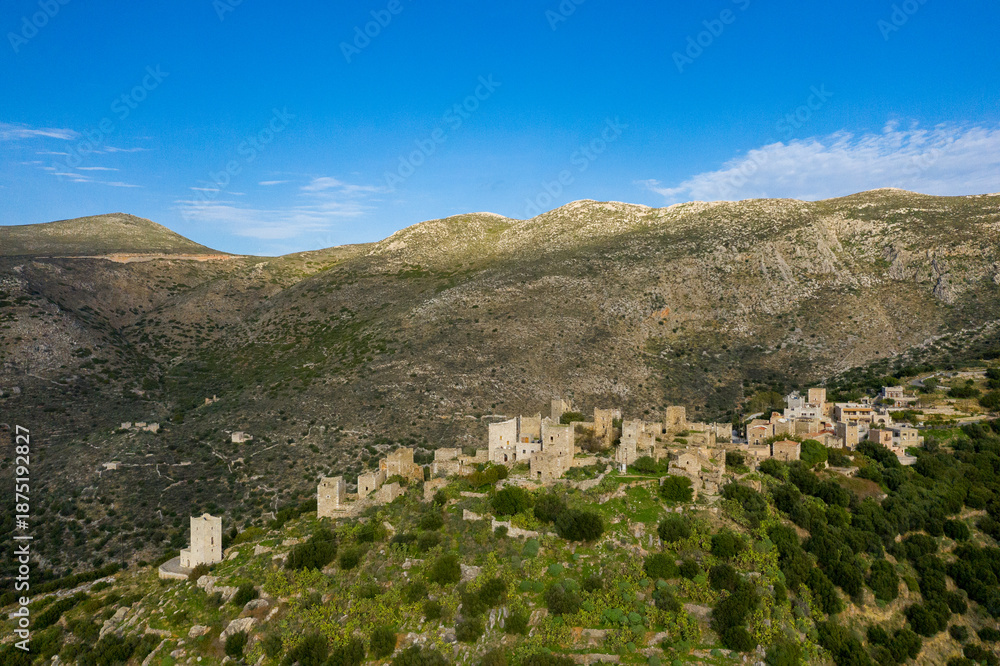Fototapeta premium Aerial view of the historic stone village of Vathia perched on a lush hillside, surrounded by rugged mountains and clear blue sky. The old towers and clustered buildings create a striking contrast