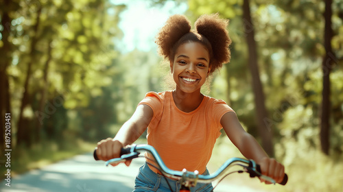 Smiling Afro-American teen girl riding bike on empty road through green trees in daylight