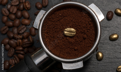 Espresso portafilter filled with ground coffee featuring a single golden coffee bean, surrounded by roasted and gold-colored beans on a dark stone background.