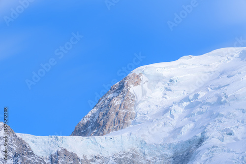 A striking view of Mont Blanc du Tacul reveals rugged rock faces emerging from pristine glacier ice under a vivid blue sky, with crisp sunlight accentuating the alpine textures.