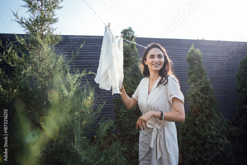 Wallpaper Mural Smiling woman with long dark hair hanging freshly washed clothes on a clothesline in a sunny garden surrounded by lush greenery, enjoying a peaceful outdoor moment Torontodigital.ca