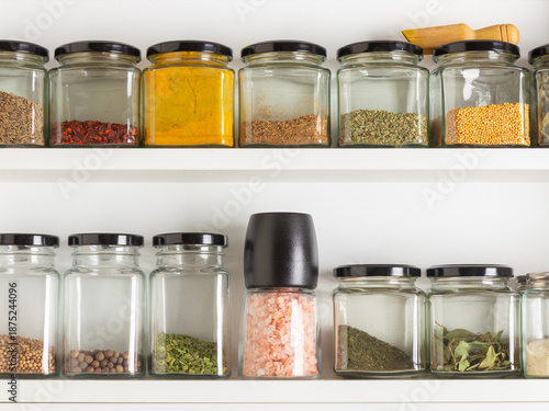 Two white shelf display an organized collection of glass jar filled with various spice and herb, suggesting a tidy kitchen, a passion for cooking, and practical domestic organization.