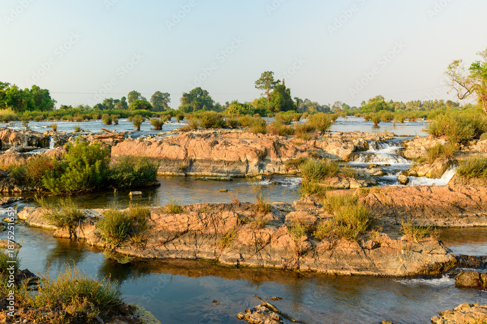 Fototapeta premium Soft morning sunlight highlights layered stone, gentle rapids, and scattered green vegetation at Tat Somphamit Waterfalls, also known as Li Phi Falls, in Champasak Province, southern Laos. The dry