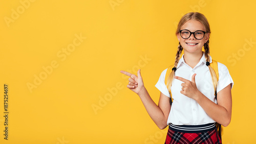 Canvas Print Smiling cute smart adolescent blonde hemale pupil in glasses shows fingers to empty space, isolated on yellow background