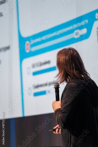 A speaker stands with a microphone facing a large screen with blue graphics. The scene suggests a presentation at a modern conference.
