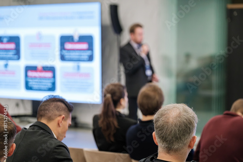 Audience attends professional presentation in conference room. Speaker conveys information on screen. Attentive listeners focus on charts and slides. Modern setting with glass walls 