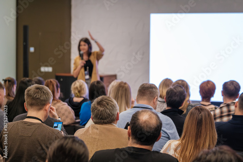 Speaker presents to a busy audience during a conference. Attentive listeners focus on the speaker while a large screen glows in the background.