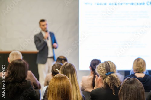 Presenter speaks to attentive audience in bright conference room. Large projection screen displays text. Attendees listen, take notes, and engage with talk in professional setting.