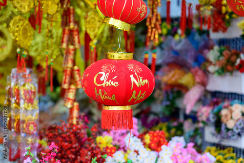 A vibrant red lantern decorated with gold calligraphy and fireworks motifs hangs among festive decorations at a market in Ben Tre, Mekong Delta, southern Vietnam. The scene features rich textures