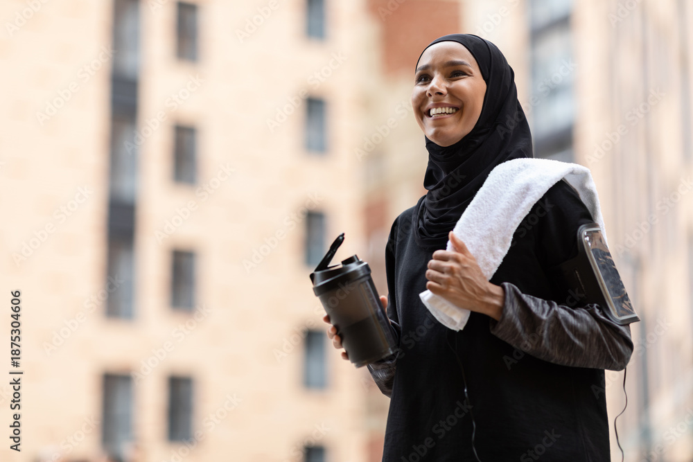 Fototapeta premium Glad young muslim lady in hijab and sportswear with phone on shoulder with cocktail cup and towel at street on building background. Outdoor workout, jogging in city, body care, ad and sports offer