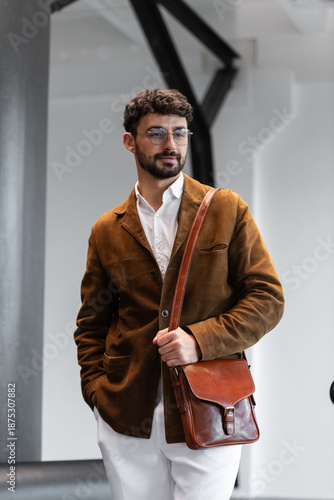 Portrait of a stylish man in a brown corduroy blazer and white shirt holding a cognac leather messenger bag in a minimalist studio.
