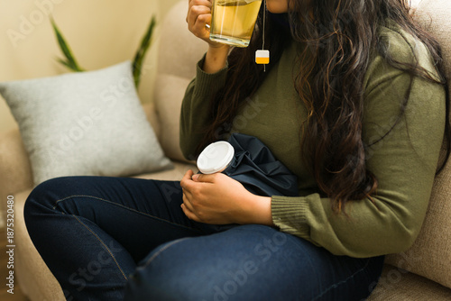 Fotografie Woman feeling menstrual pain, drinking a hot herbal tea and applying a warm comp