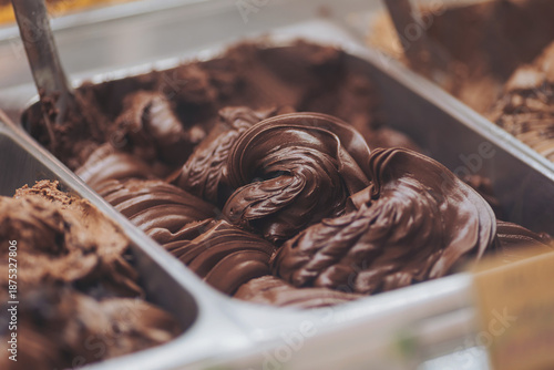 Chocolate gelato fills a large metal container in a dessert shop. The rich texture attracts attention as two scooping spoons rest nearby. Customers enjoy the warm afternoon air