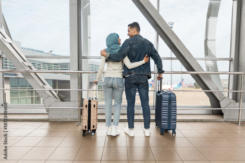 Fototapeta premium Romantic Travel. Loving Muslim Couple Standing With Suitcases Near Window In Airport, Happy Islamic Spouses Embracing And Smiling To Each Other, Ready To Honeymoon Trip, Rear View With Copy Space