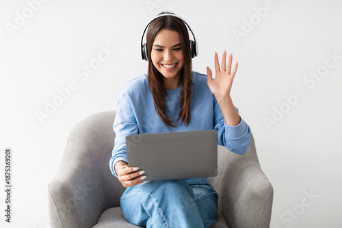 Wallpaper Mural Distant virtual communication concept. Pretty young lady in headphones waving at laptop screen, sitting in armchair, making video call, chatting online on white studio background Torontodigital.ca