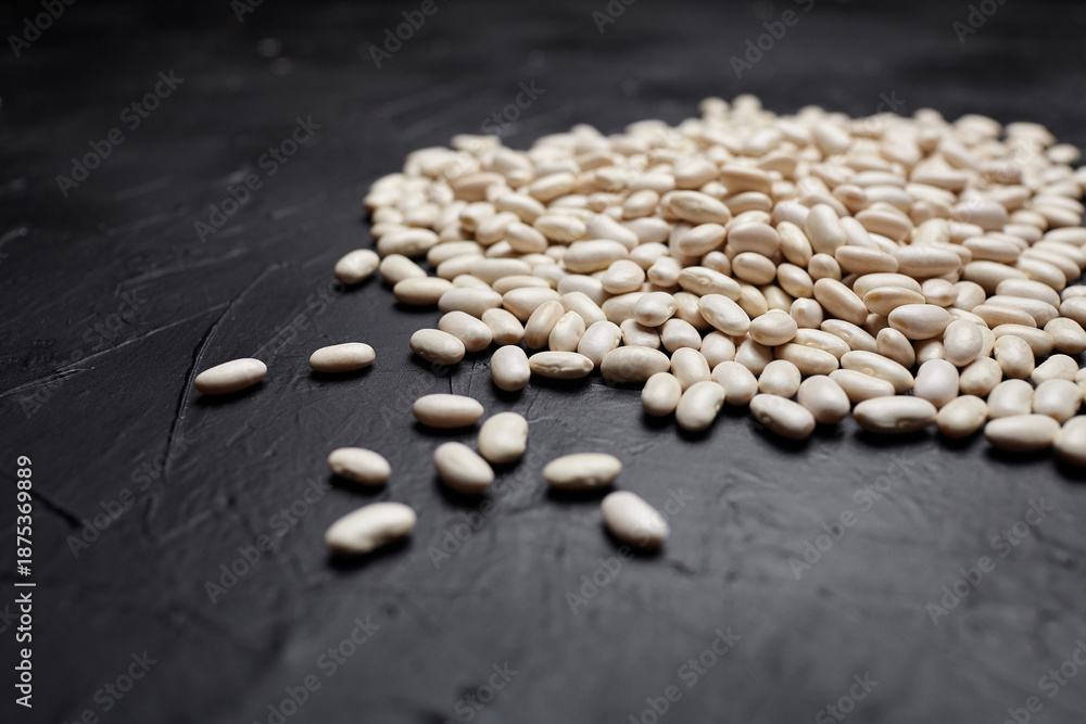 Fototapeta premium White beans scattered on a dark textured surface, showcasing their smooth, creamy appearance and natural sheen, perfect for culinary and health-related concepts