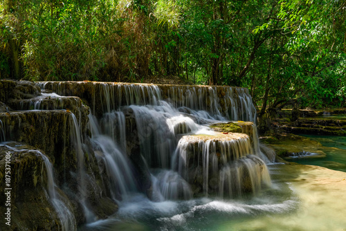 Gentle streams of water flow over tiered limestone formations surrounded by dense green forest at Kuang Si Falls in northern Laos. Sunlight highlights the cascading water and textured rocks in this