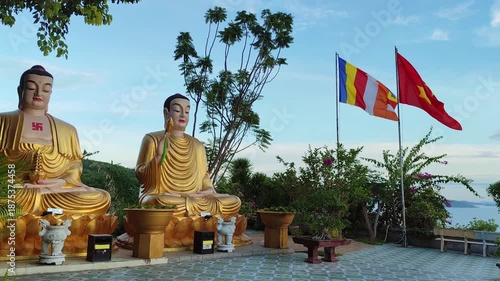 Two serene sitting Buddha statues with colorful Buddhist flags at Chua Da Bao Pagoda in Nha Trang, Vietnam. Peaceful spiritual scene in a traditional temple surrounded by nature and mountain views.