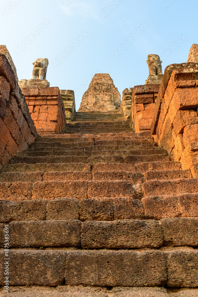 Fototapeta premium Low-angle view of ancient stone steps ascending toward the weathered central tower of Pre Rup temple in Cambodia, flanked by eroded guardian statues. The scene is bathed in warm late afternoon light