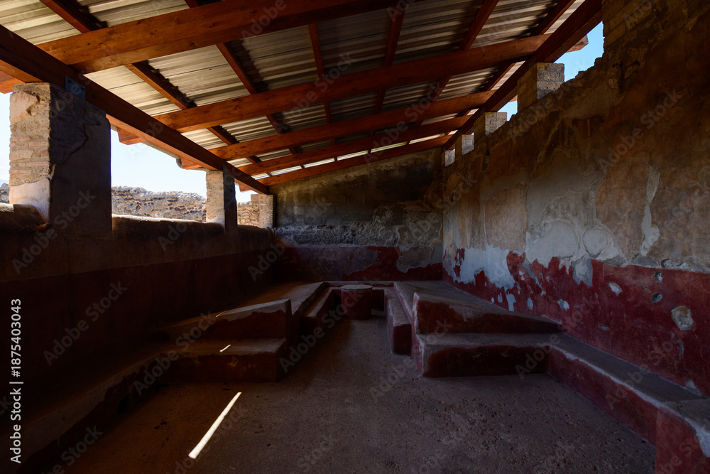 Fototapeta premium Stone benches line an ancient Roman dining room with faded red frescoes and weathered plaster in the House of the Lararium of Achilles, Pompeii, Italy. Diffused daylight enters through open windows