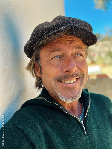 Fotografía Smiling rugged man with woolen newsboy cap and stubble beard in green woolen sweater standing outside by white wall of spanish house in countryside