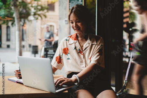 Konstfotografi Smiling Asian woman works on laptop near window, combining casual style with digital fluency, expressing confidence and ease in tech-driven freelance or study lifestyle