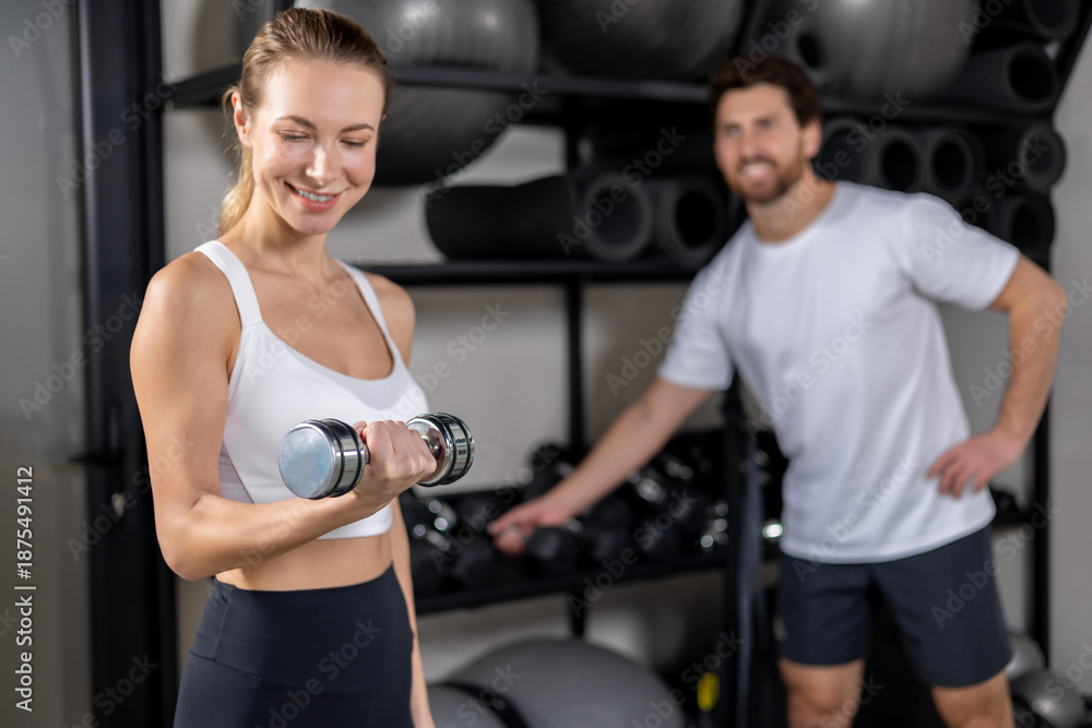 Fototapeta premium Woman in white top and black leggings exercising with the dumbbells in the gym