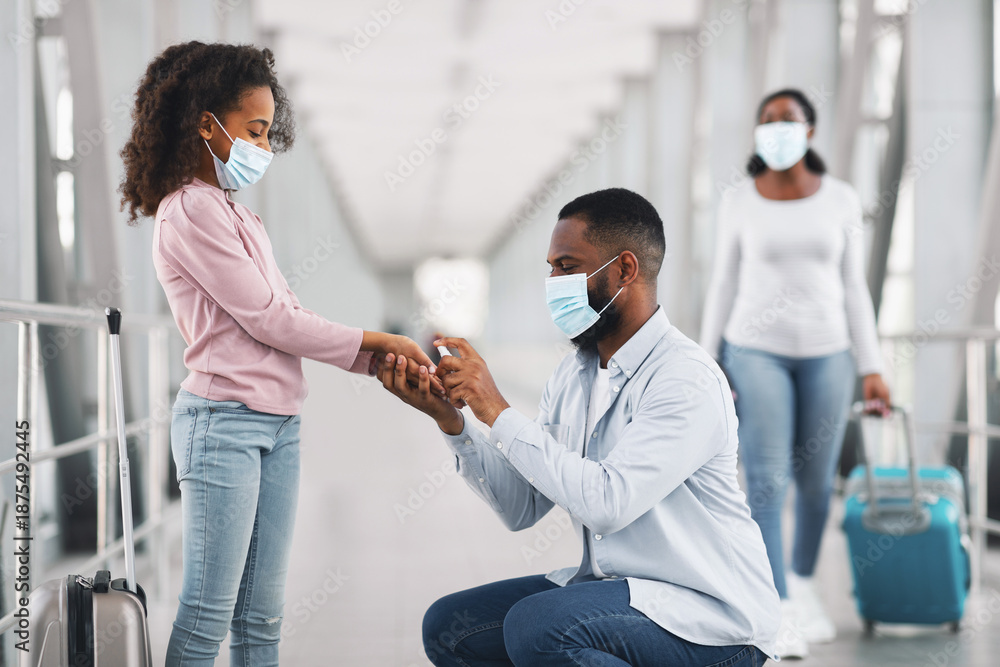Fototapeta premium Health Care And New Normal Protection Concept. Black Father Spraying Sanitizer On Daughter's Hands Cleaning Arms In The Airport Terminal Indoors, Wearing Protective Medical Masks For Covid Prevention