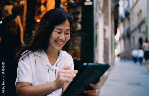 Slika na platnu Asian woman smiling while using digital tablet with stylus, showing delight in creative process, digital autonomy and confident freelance mindset in mobile work