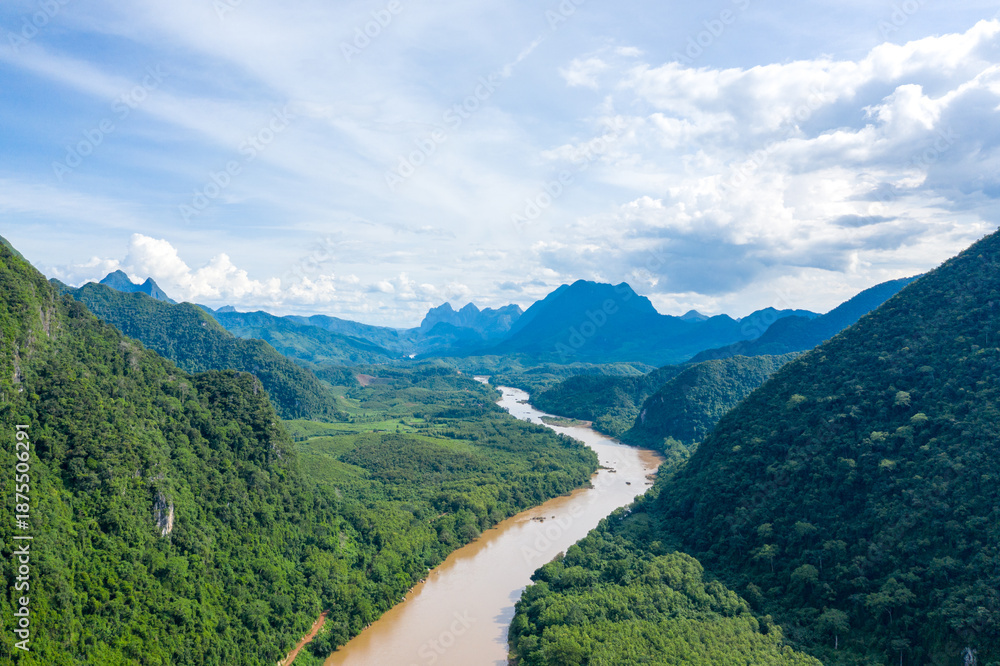 Obraz premium Wide aerial perspective of the Nam Ou river winding through lush green hills and forested mountains near Muang Ngoi in northern Laos. Bright blue sky and dramatic cloud formations enhance the sense of