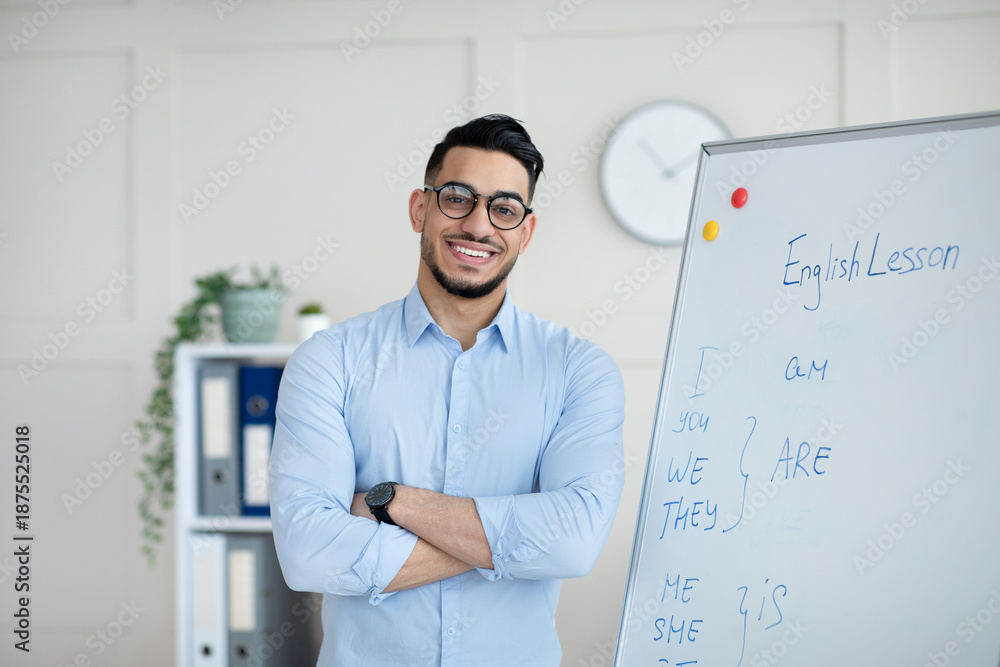 Obraz premium Distance learning. Happy Arab male teacher giving online English class, standing with crossed arms near blackboard, smiling at camera in home office. College tutor explaining grammar rules to students