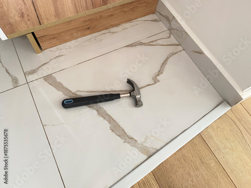 A metal hammer with a rubber handle lies on ceramic floor tiles next to a kitchen cabinet and wooden flooring in a residential interior.